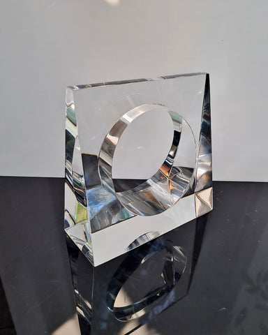 The Liquid Lucite Wedge Bangle Bracelet by Nik Spruill, a clear, geometric masterpiece with a distinctive circular opening at its center, is displayed on a reflective black surface against a neutral backdrop, radiating an elegance akin to sophisticated jewelry.