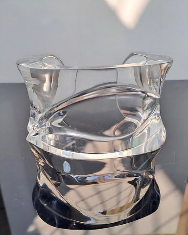 A Luminous Lucite Cuff Bangle Bracelet by Nik Spruill, showcasing intricately folded glass with ripple wave details and smooth surfaces that radiate luminous reflections, set on a black surface with a white wall background.