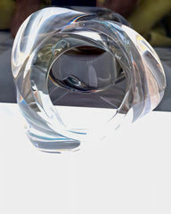 A clear, hand-sculpted abstract glass sculpture with curved and intertwined shapes, reminiscent of the Liquid Lucite Asymmetrical Bangle Bracelet by Nik Spruill, displayed on a reflective surface.
