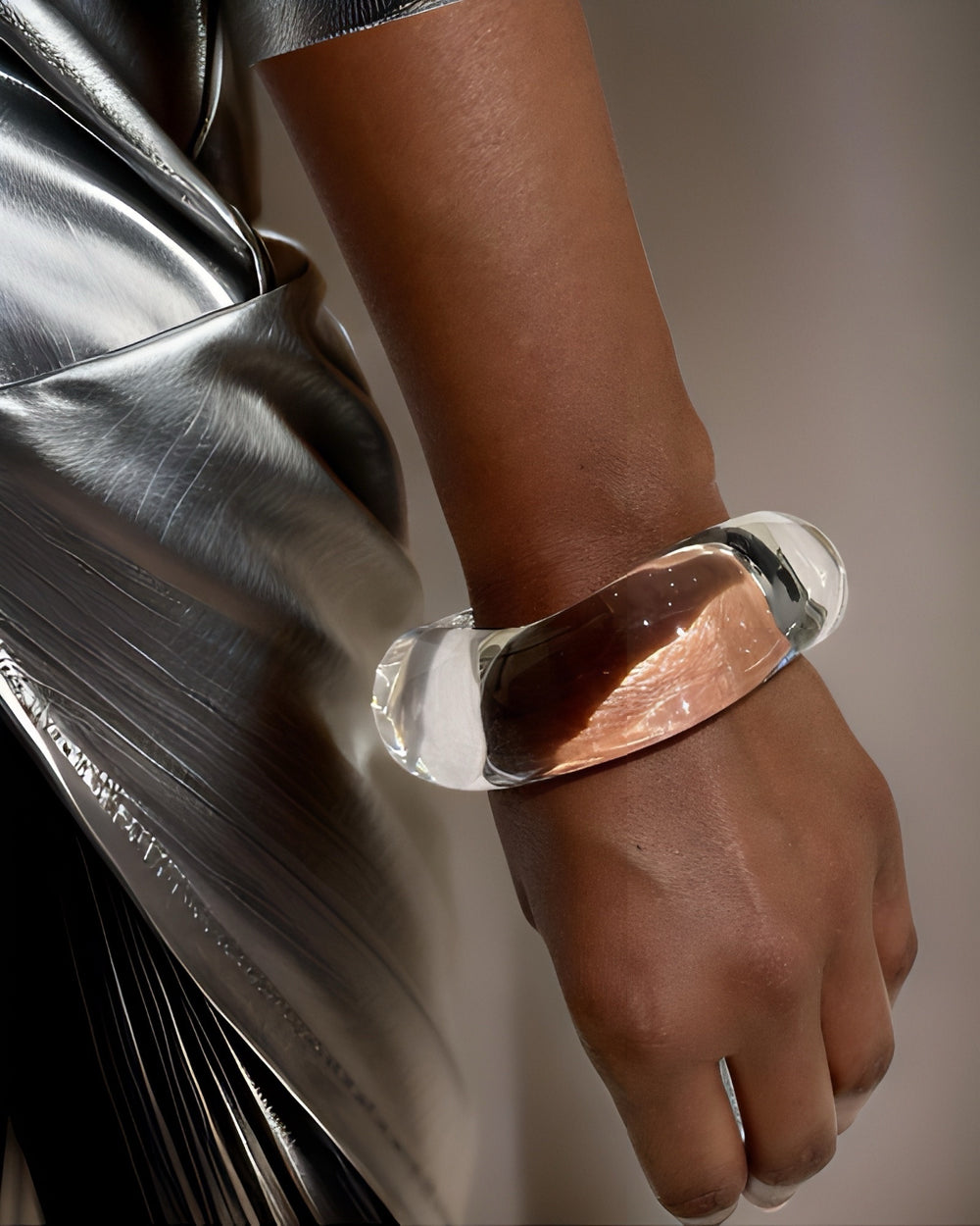 Close-up of a person's wrist adorned with a clear, chunky Lucite Stackable Circle Bangle Bracelet by Nik Spruill. The person is dressed in a metallic silver outfit. The lightweight design adds elegance, while the background remains neutral.