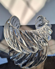 Wiggle Lucite Cuff Bracelet by Nik Spruill features a clear glass-like design with an intricate spiral pattern, positioned on a reflective black surface, casting a soft shadow that enhances its stunning statement as an accessory.