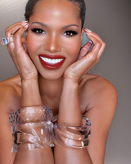 A smiling person with dark hair pulled back is wearing clear plastic bracelets, including the Nik Spruill Liquid Lucite Wave Cuff Bracelet and a large ring, and posing with their hands near their face.