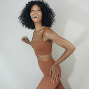 A person with curly hair, wearing a caramel SHIVAYA CROP TOP from Nik Spruill and a brown athletic outfit, smiles and poses against a plain background.